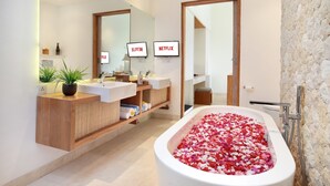 Un baño moderno con lavamanos dobles, un espejo grande, una bañera llena de pétalos de rosa y dos pantallas incorporadas mostrando el logotipo de Netflix.