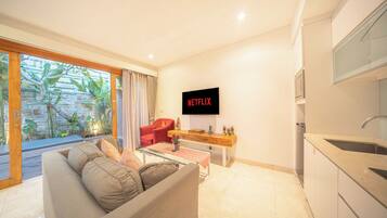 A modern living room with a sofa, a coffee table, a red chair, a kitchen area with a sink, and a wall-mounted TV displaying "NETFLIX".