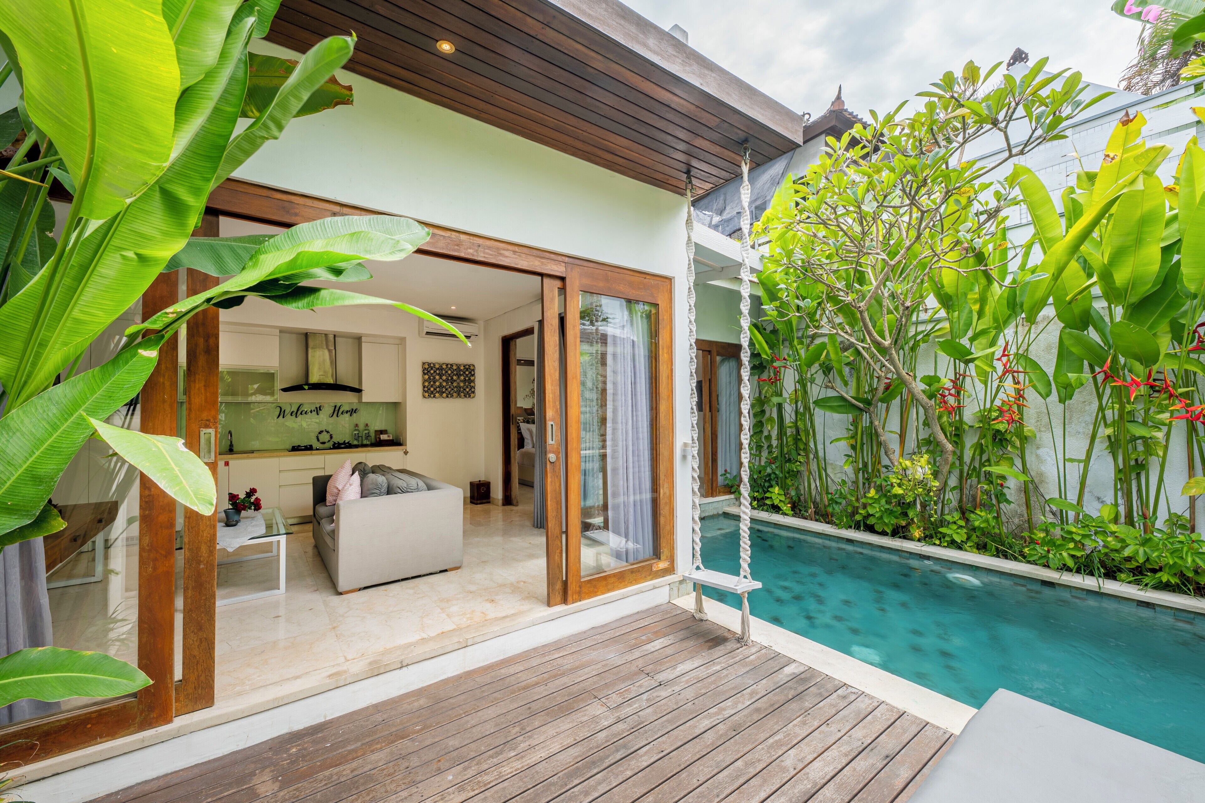 Una casa moderna con piscina, un salón con sofá y mesa de centro, y una cocina visible a través del ventanal.
