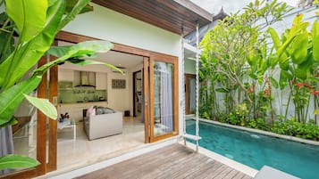 Uma casa moderna com piscina, uma sala de estar com sofá e mesa de centro, e uma cozinha visível pela porta de vidro.