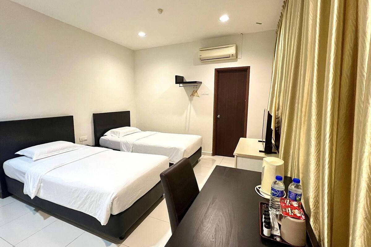 A hotel room with two beds, a desk with a lamp, a chair, and a small table with water bottles and snacks.