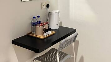 Une petite pièce moderne, avec un bureau noir, une chaise, une bouilloire blanche et un plateau contenant des bouteilles et une tasse.