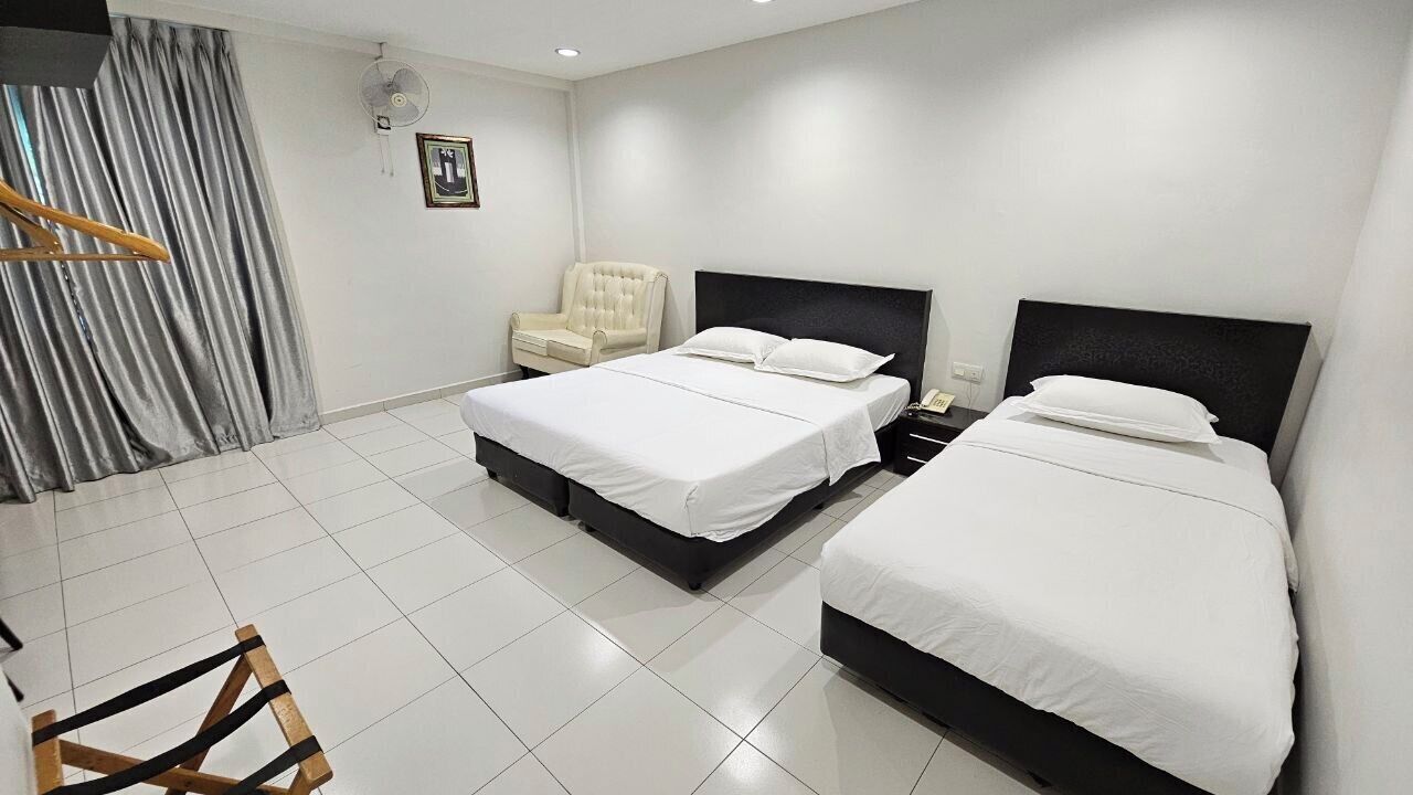 A hotel room with two beds, a chair, a bedside table, a fan, and a curtain.