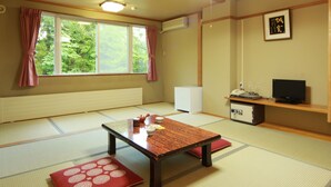 Ein traditioneller japanischer Raum mit Tatami-Boden, einem niedrigen Tisch mit Kissen, einem Fernseher und einem Fenster mit Vorhängen.