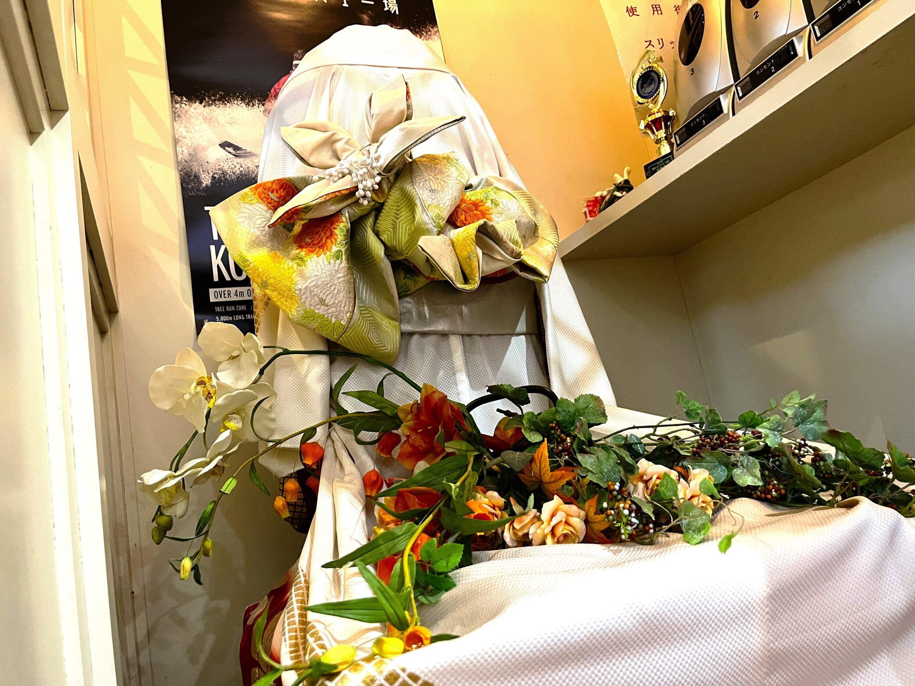 A mannequin dressed in a white garment with a large bow, surrounded by various flowers and plants.