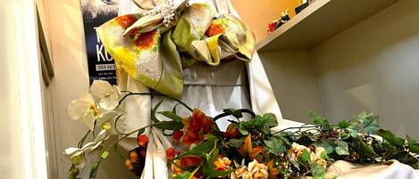 A mannequin dressed in a white garment with a large bow, surrounded by various flowers and plants.