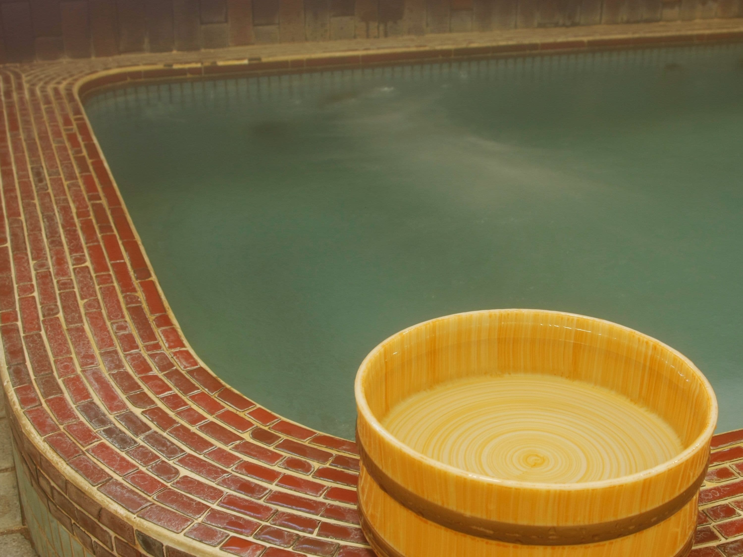 Una piscina con borde de ladrillo y una bañera de madera al frente.