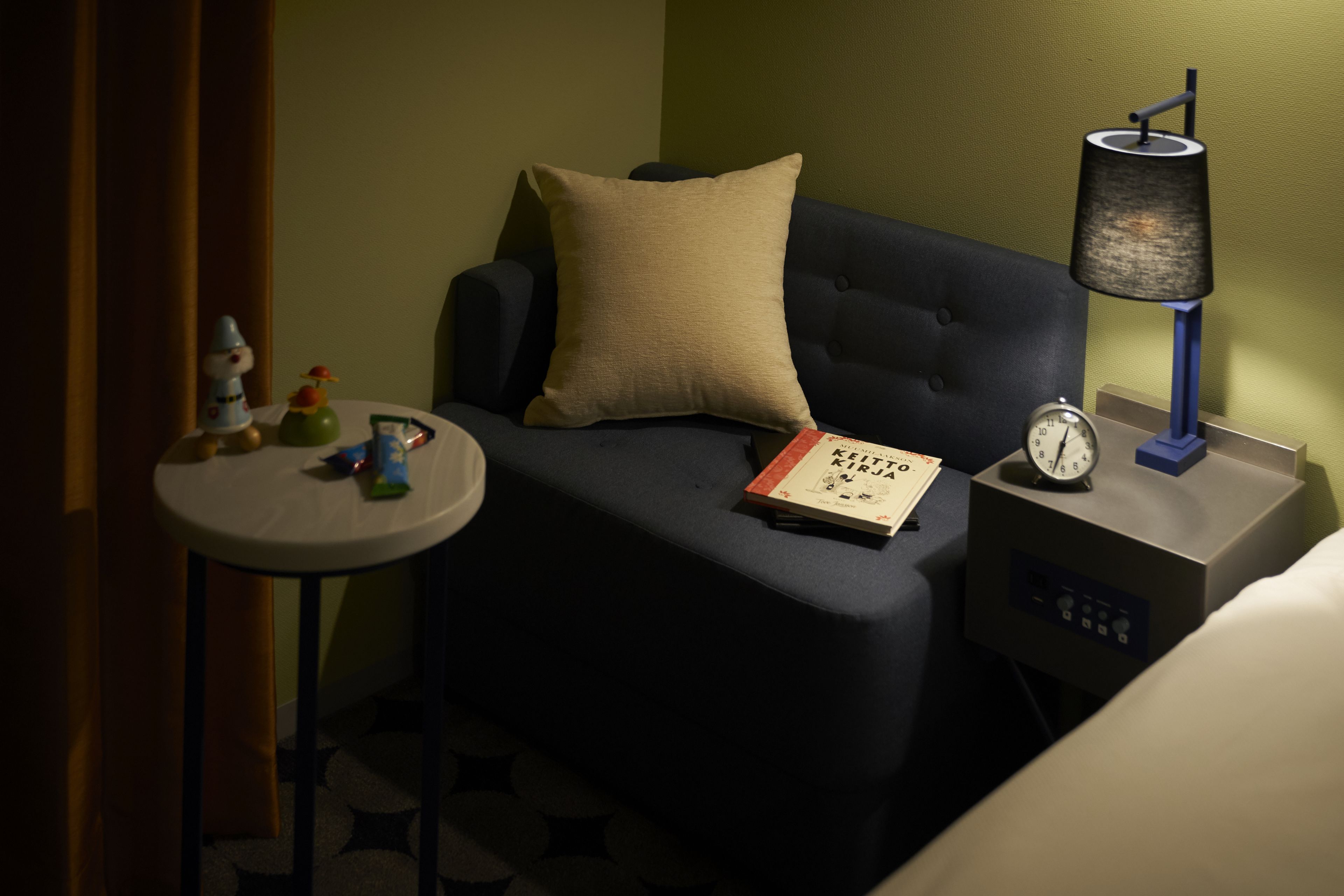 Een hotelkamer met een bed, een nachtkastje met een lamp, een klok en een boek.