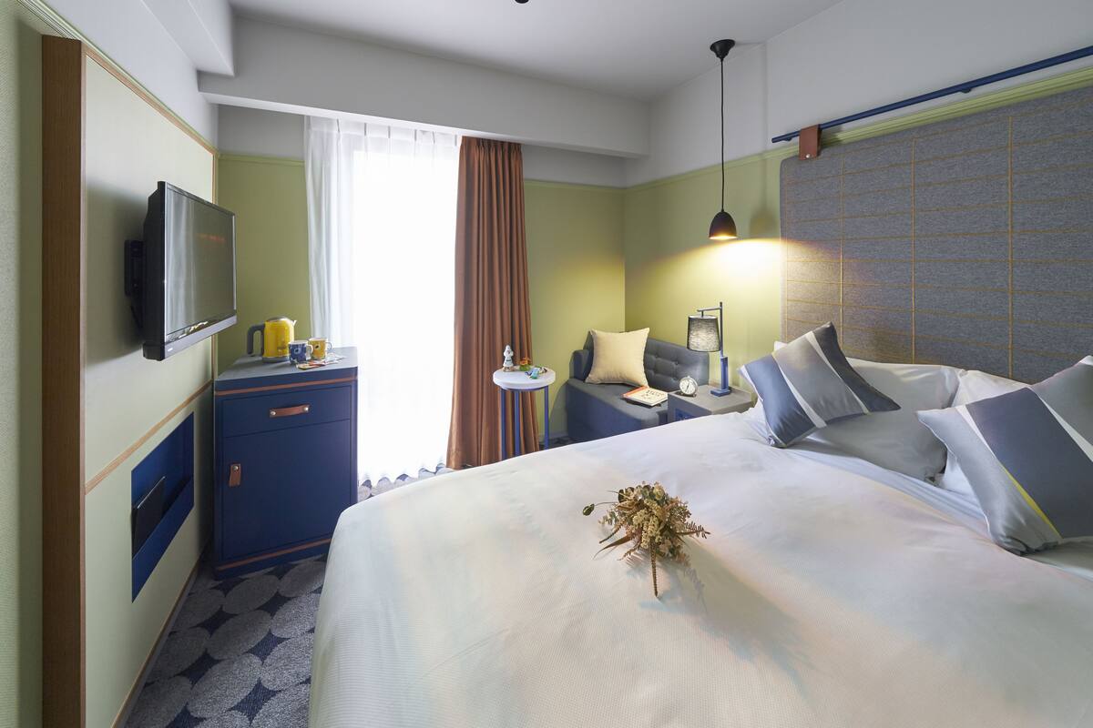 Habitación de hotel con cama, mesitas de noche, televisión, una mesita pequeña con hervidor y ventana con cortinas.