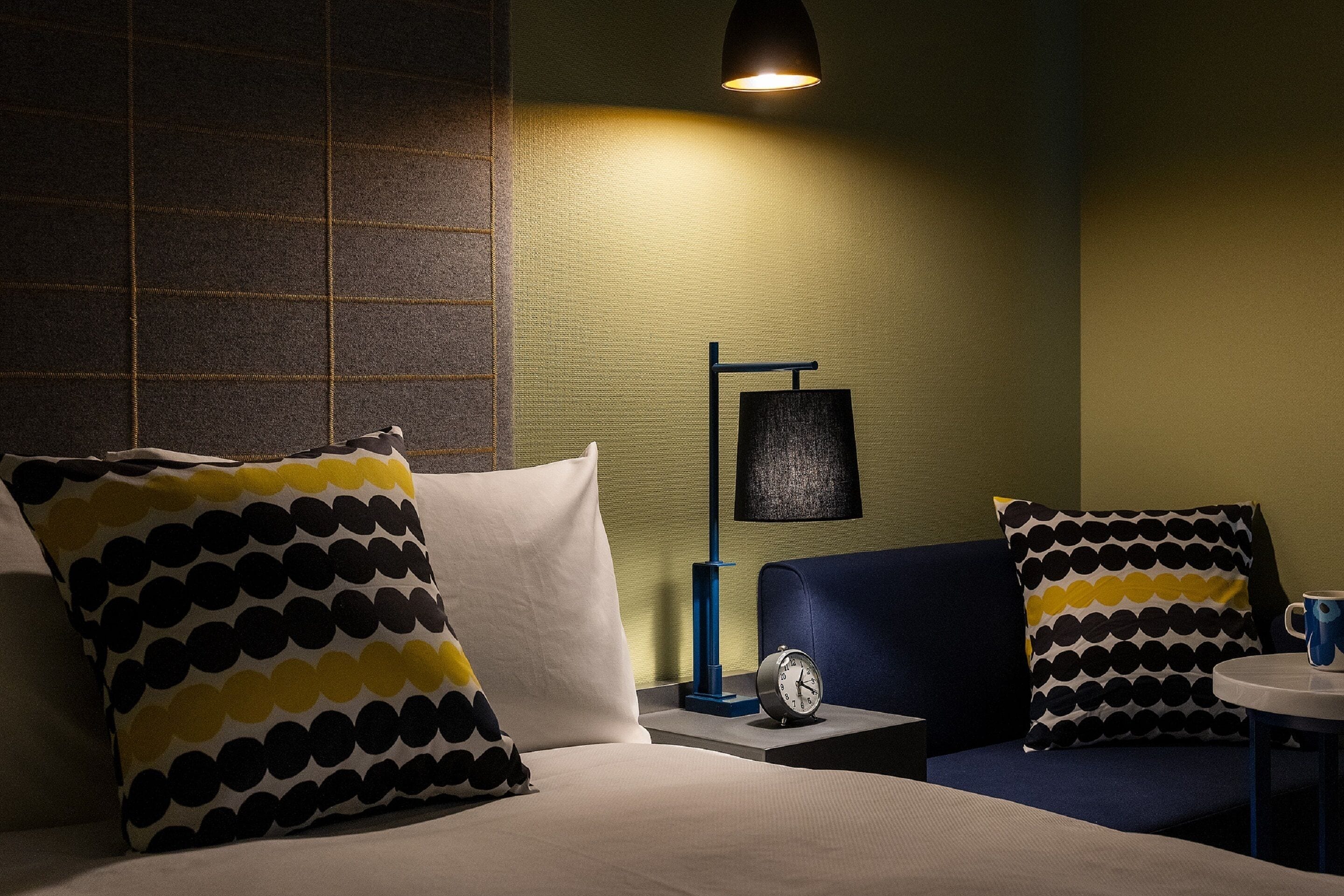 Une chambre d’hôtel avec un lit, deux tables de chevet, une lampe, une horloge et une petite table sur laquelle se trouve une tasse.