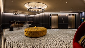 Een moderne hotel lobby met een vloer in geometrisch patroon, een centrale ottoman met knoopjes, een kroonluchter en een lift.