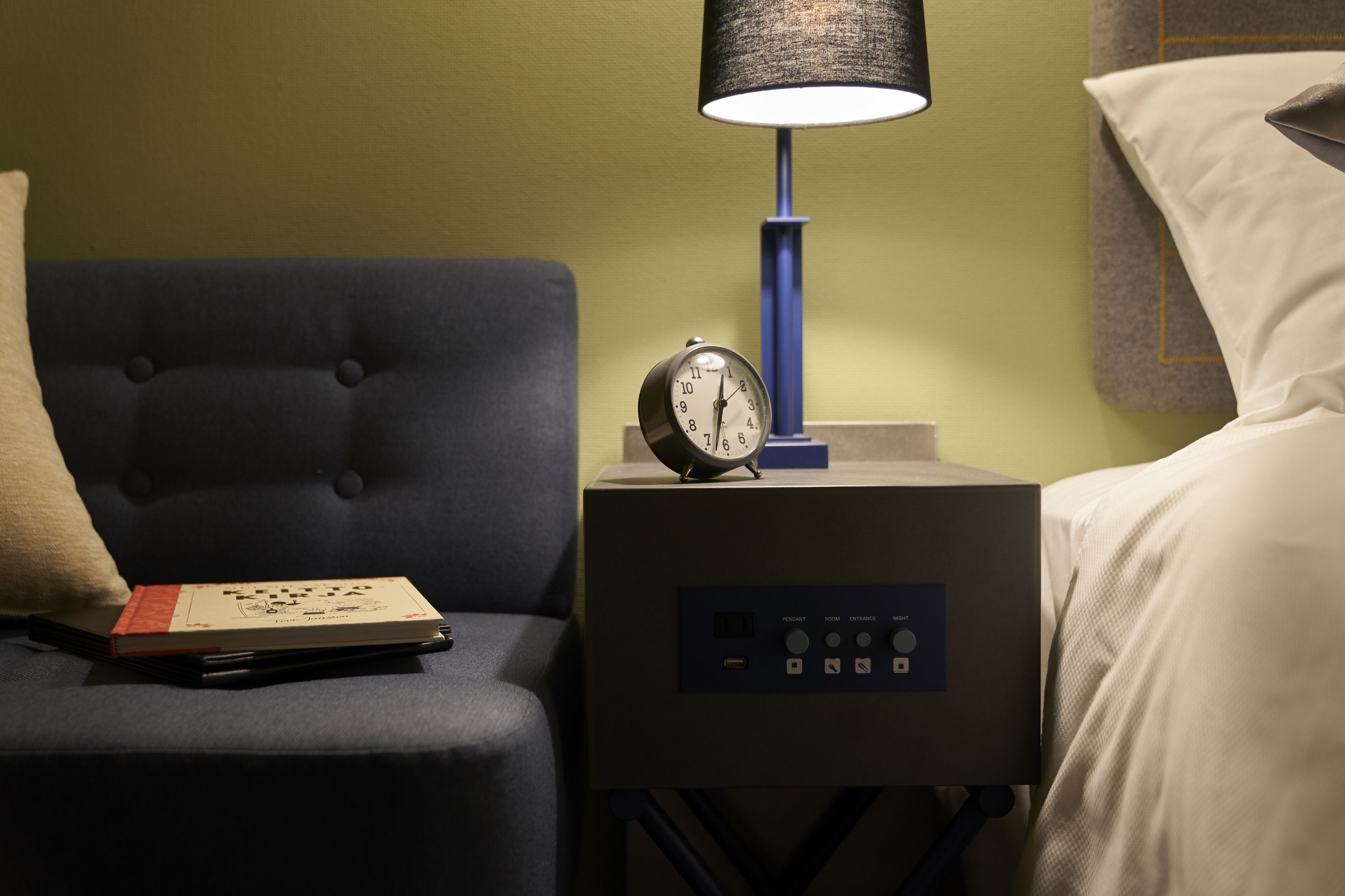 A neatly made bed with white linens, a bedside table with a clock, a lamp, and a book on a dark blue upholstered chair.