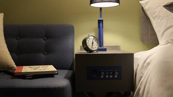 A neatly made bed with white linens, a bedside table with a clock, a lamp, and a book on a dark blue upholstered chair.