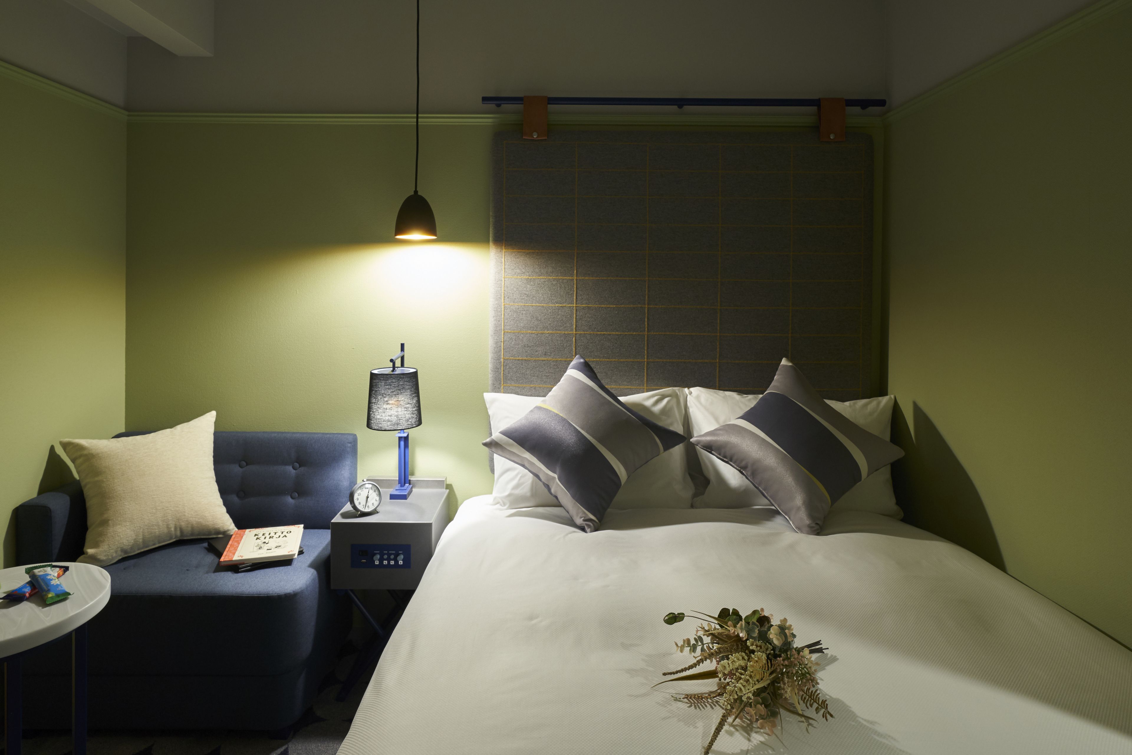 Una habitación de hotel con una cama, una mesita de noche con lámpara, una pequeña zona de estar con sillón y una pared con diseño de cuadrícula.