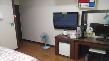 Ein Hotelzimmer mit einem Bett, einem Schreibtisch mit Computer, einem kleinen Kühlschrank, einem Ventilator und einem Feuerlöscher.