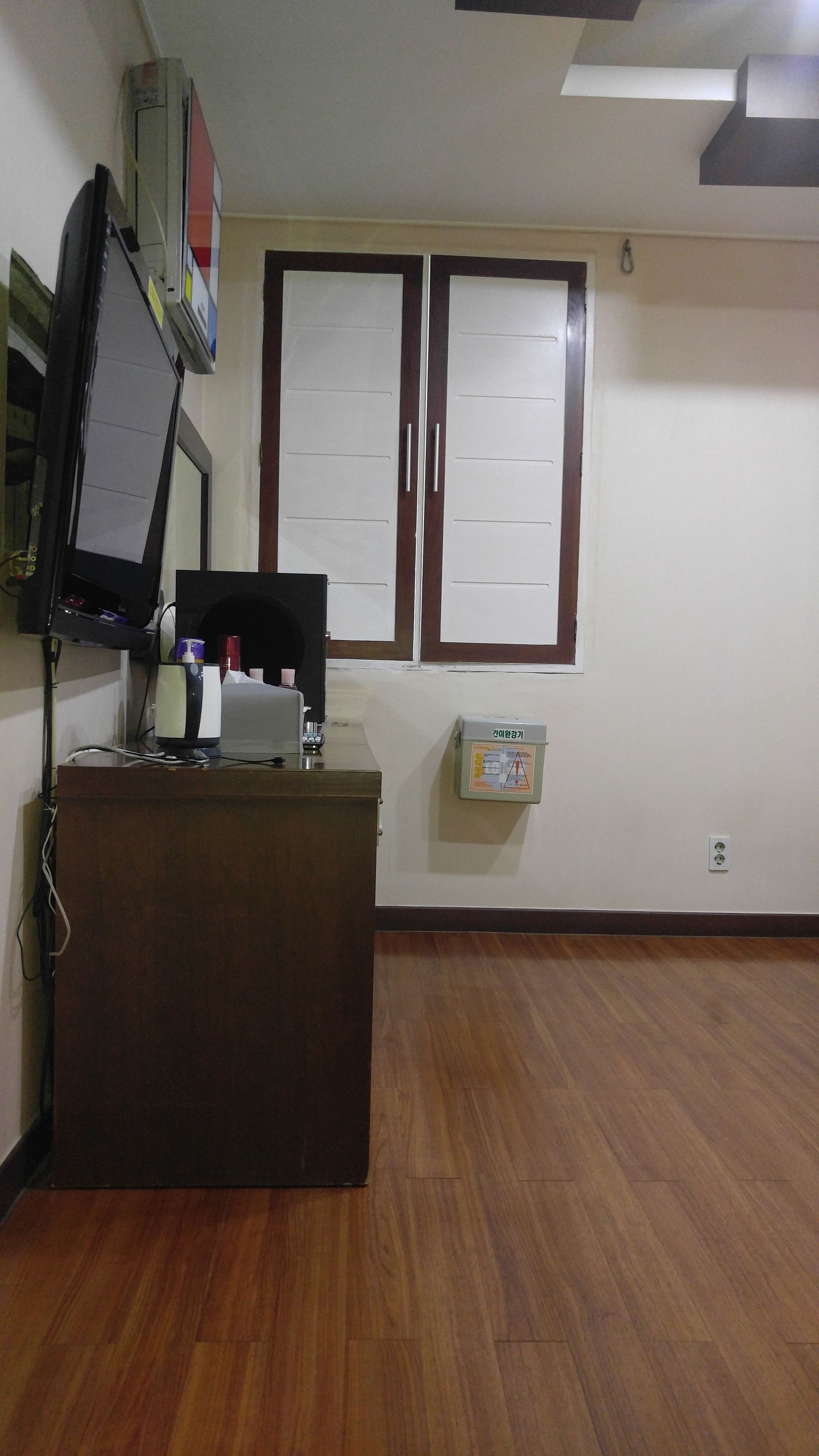 A room with a wooden desk, a flat-screen TV, two windows with blinds, a wall-mounted electrical box, and a ceiling vent.
