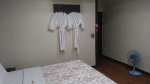 A hotel room with a bed, a patterned bedspread, two white robes hanging on the wall, a blue standing fan, and a fire alarm on the wall. - Odyssey  Hotel (Chungju)