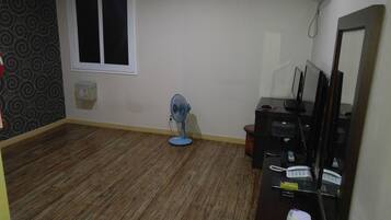 Una habitación con piso de madera, un ventilador de pie, un escritorio con computadora y un teléfono.