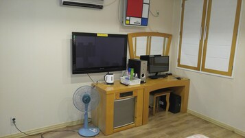 Espaço de escritório compacto com escrivaninha de madeira, dois monitores, um ventilador e um frigorífico pequeno.