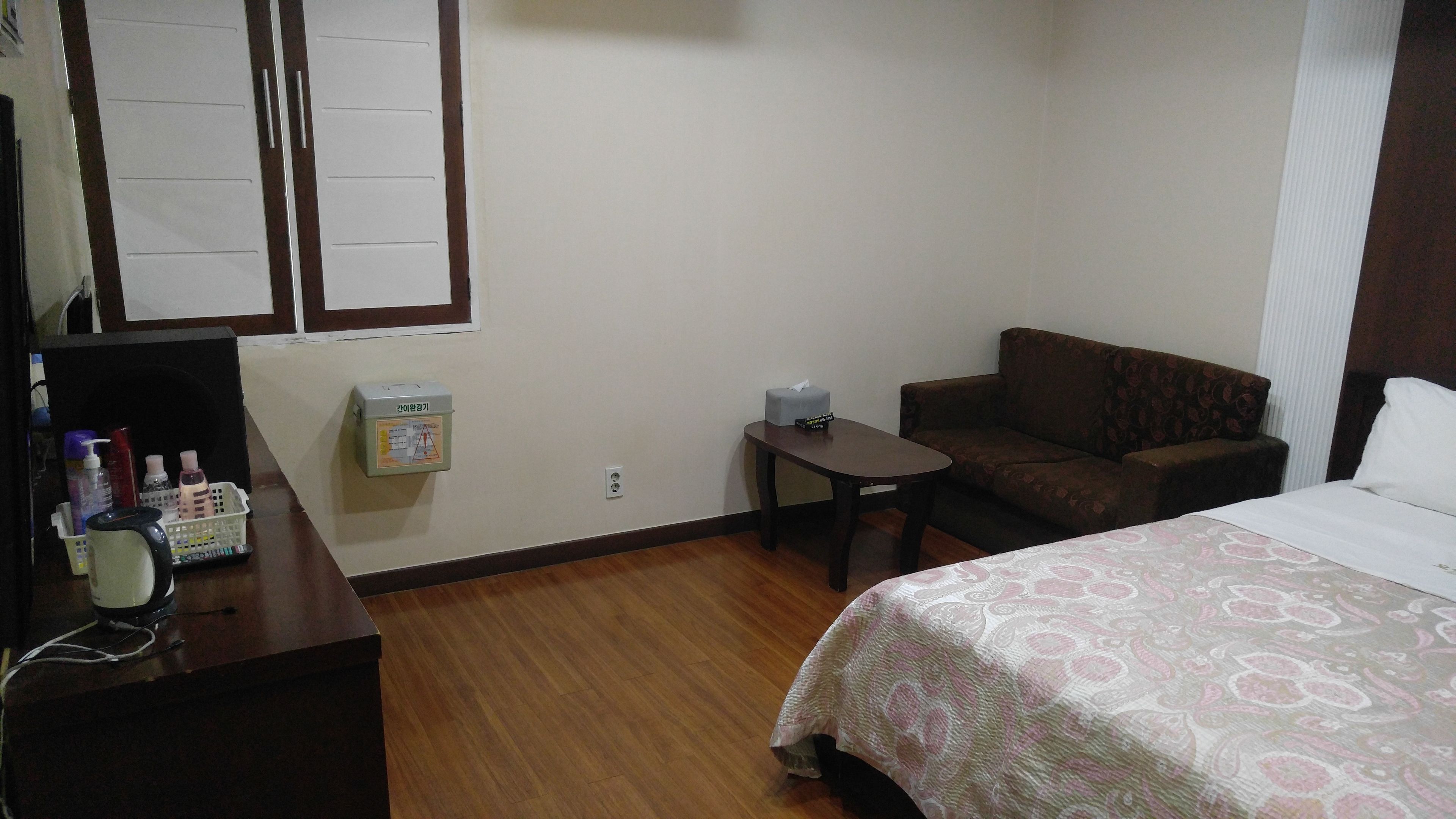 A hotel room with a bed, a sofa, a small table, a desk with a computer and toiletries, and a wall-mounted emergency box.