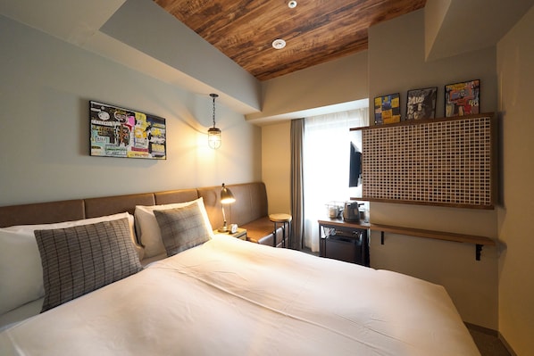 Una camera d'albergo con un letto grande, una testiera, un comodino con una lampada, una scrivania e una finestra con tende.