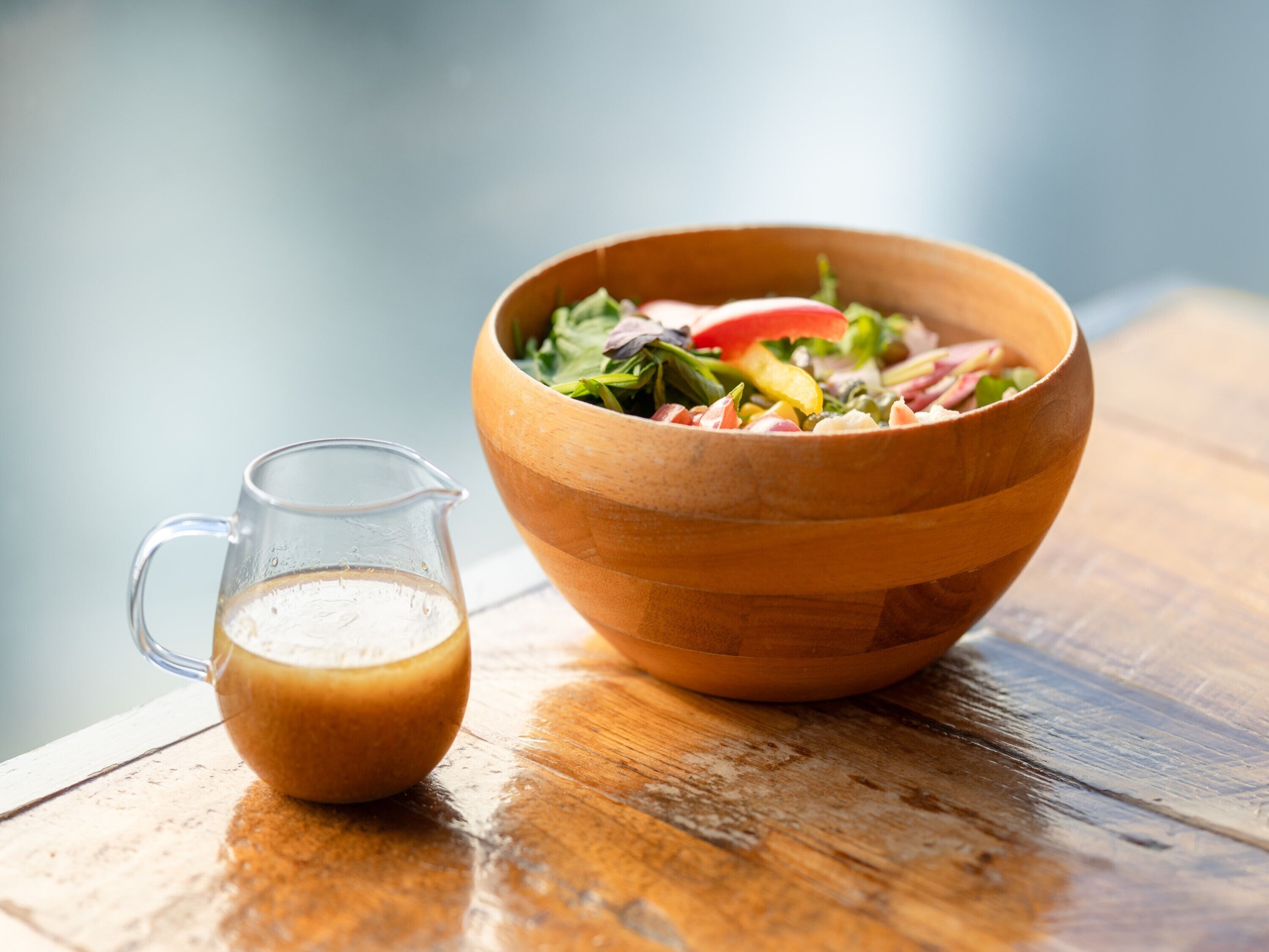 A wooden bowl filled with salad and a glass pitcher of dressing on a wooden table.