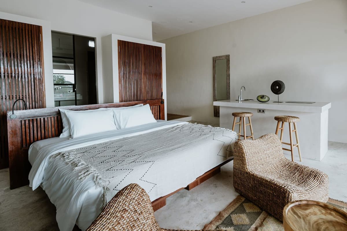 Une chambre moderne avec une tête de lit en bois, un grand lit recouvert d’une courtepointe blanche, deux fauteuils en osier, un petit bureau avec un miroir et une vue sur l’extérieur.