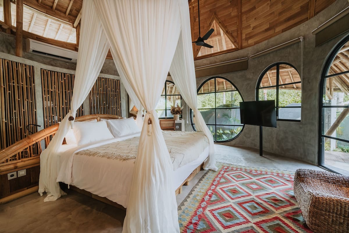 Une chambre dotée d’un lit à baldaquin, de grandes fenêtres, d’un ventilateur de plafond et d’une chaise en osier.
