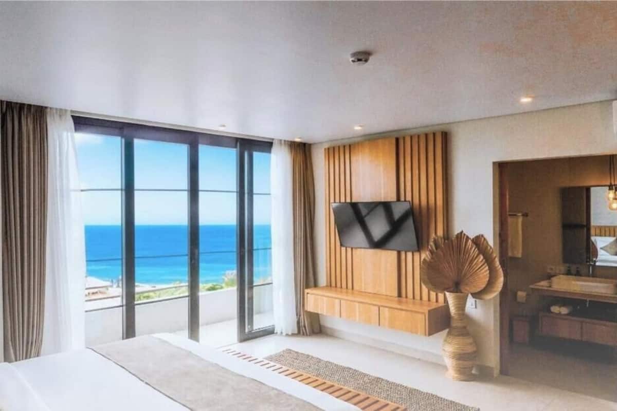 Une chambre d’hôtel moderne dotée d’une grande fenêtre, d’un téléviseur à écran plat, d’un banc en bois et d’une salle de bain visible à travers une porte ouverte.