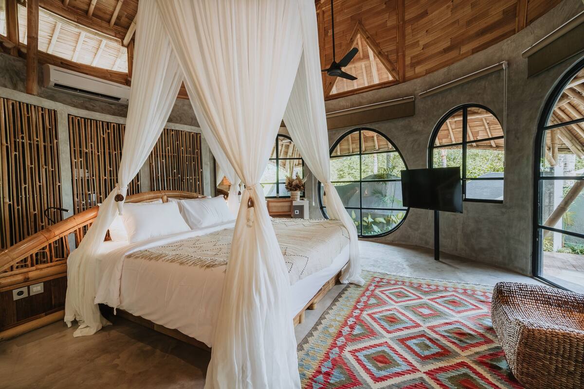 Une chambre avec un lit sous un dais, de grandes fenêtres et un ventilateur de plafond.