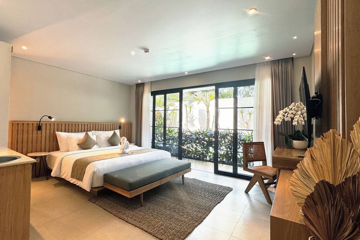 Une chambre d’hôtel moderne avec un grand lit, un petit bureau, un lavabo et un balcon agrémenté de plantes.