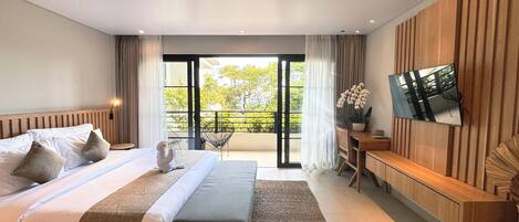 A modern hotel room with a large bed, a TV mounted on the wall, a balcony with a view of greenery, and a small desk with a chair.