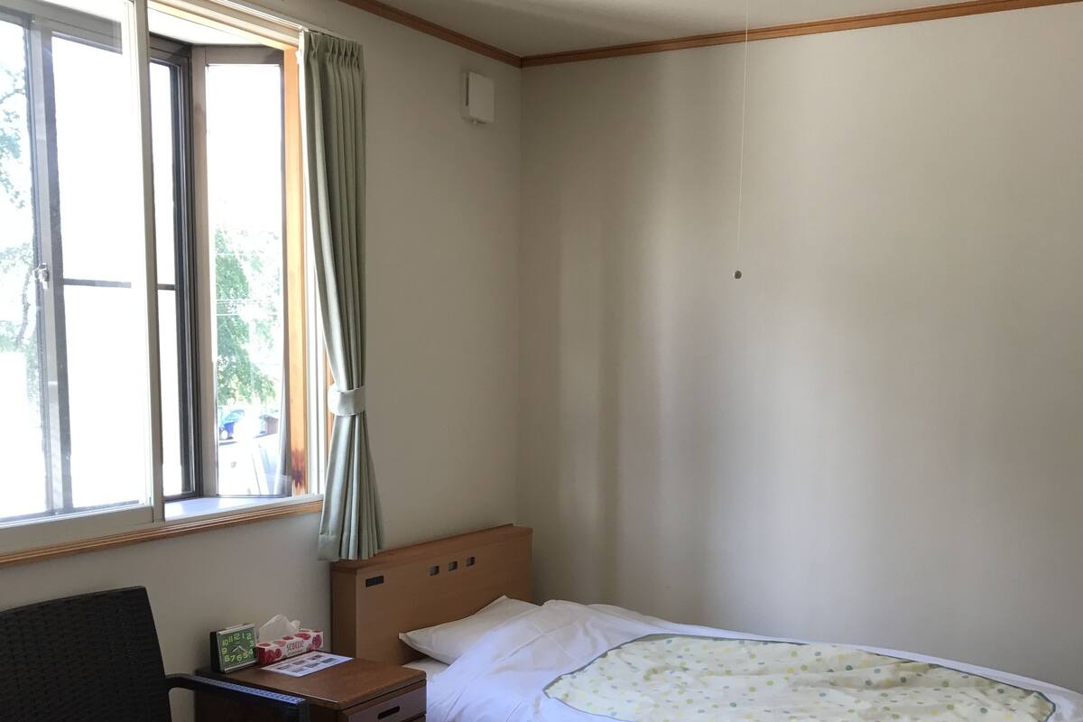 A bedroom with a single bed, a wooden desk, a chair, a waste bin, and a window with curtains.