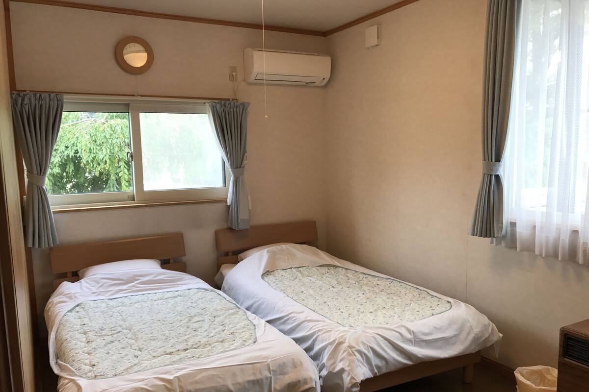 A small, clean room with two single beds, a window with curtains, an air conditioning unit, and a waste bin.