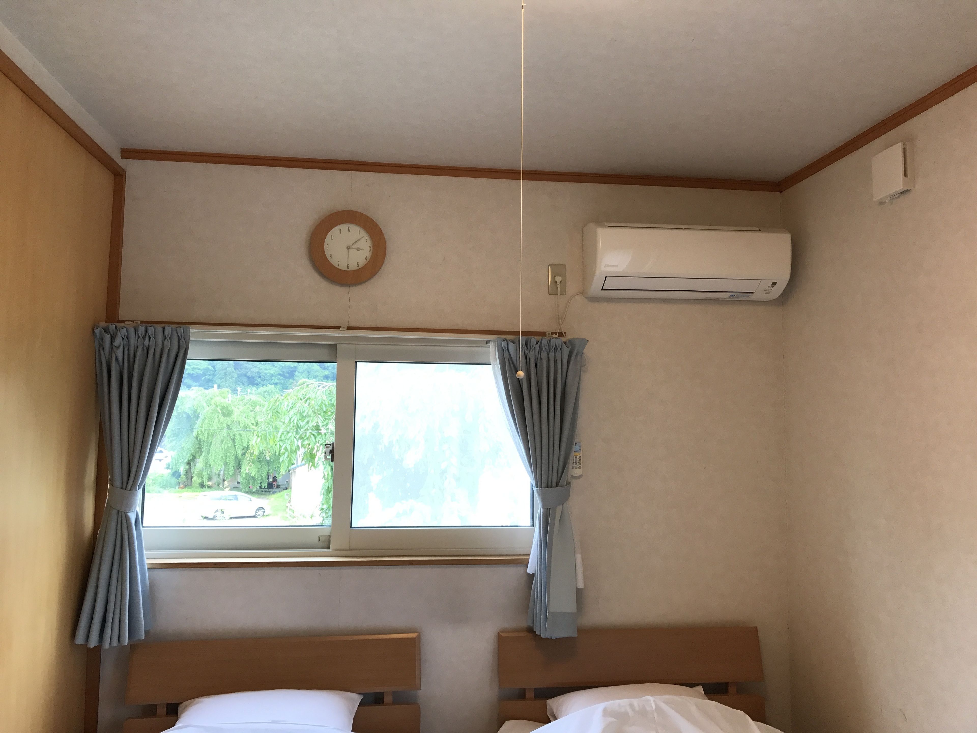 A room with two beds, a window with curtains, a wall clock, and an air conditioning unit.