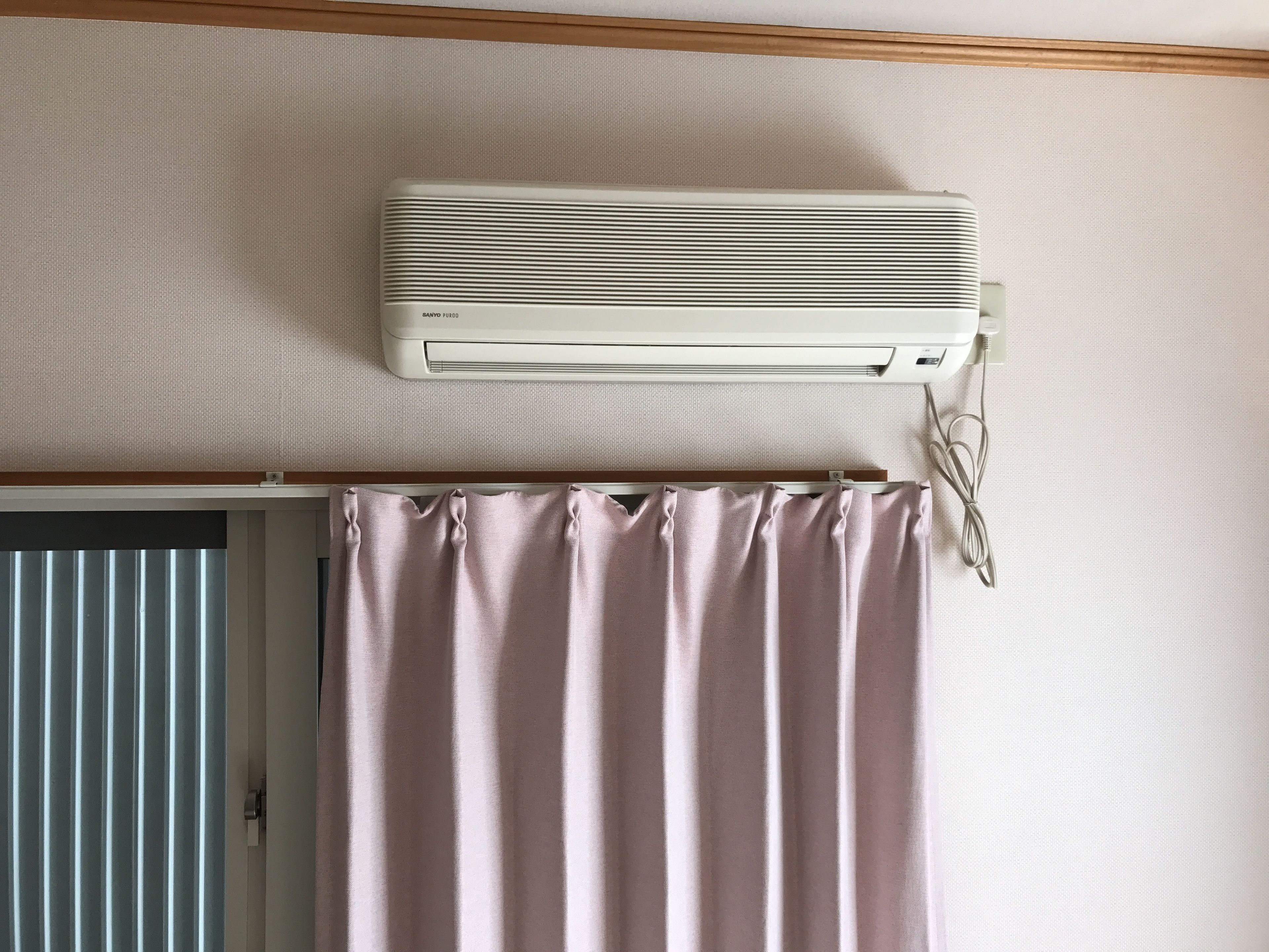 Una habitación con un aire acondicionado blanco montado en la pared, una puerta con persiana vertical y una cortina rosa.