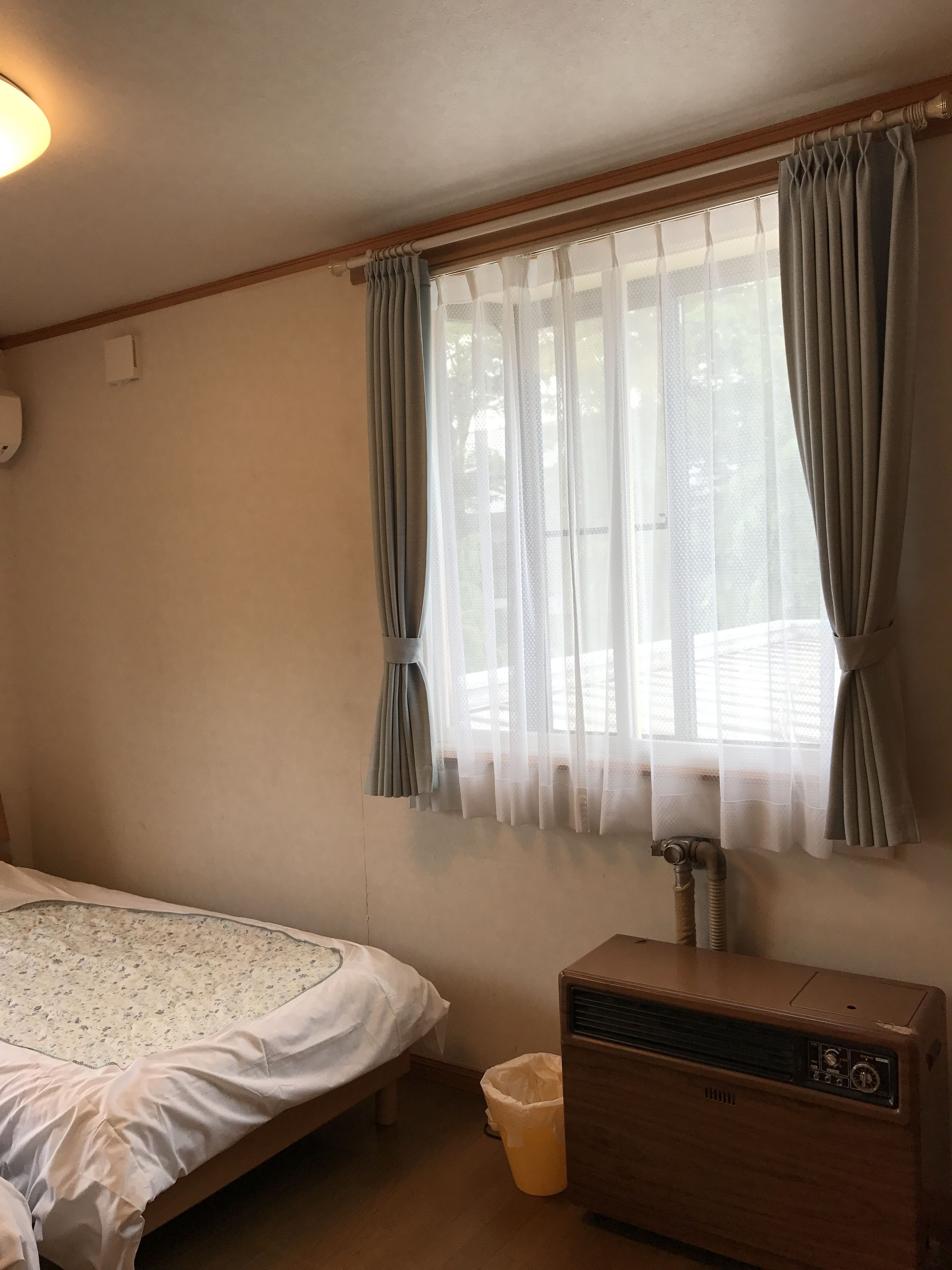Quarto com cama, radiador, janela com cortinas e luminária fixada na parede.