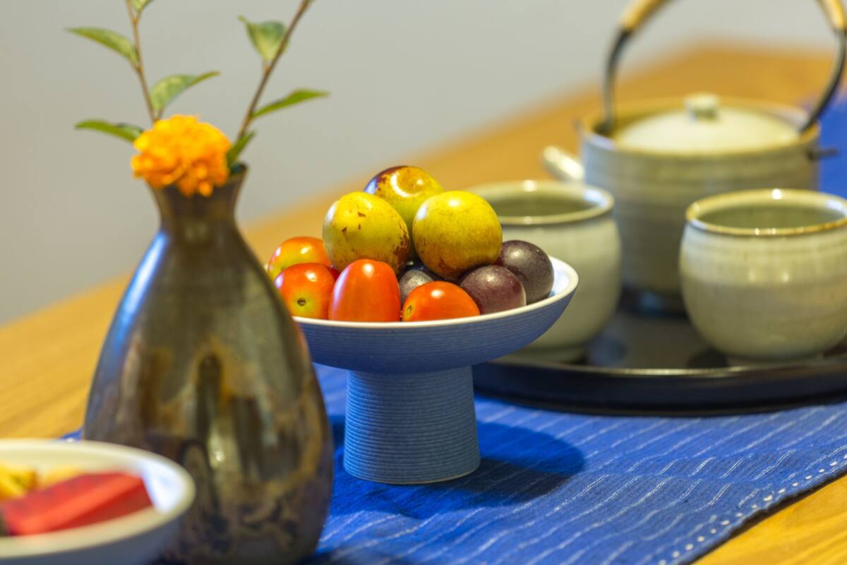 Une table à manger avec un saladier de fruits, un vase avec une fleur et une théière.
