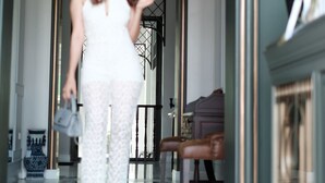 Uma mulher num macacão branco caminha por um corredor com piso de mármore, lareira e um espelho grande.