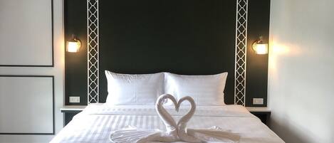Cama bem arrumada com toalha em formato de coração nos travesseiros, cabeceira estampada em preto e branco e duas luminárias fixadas na parede.