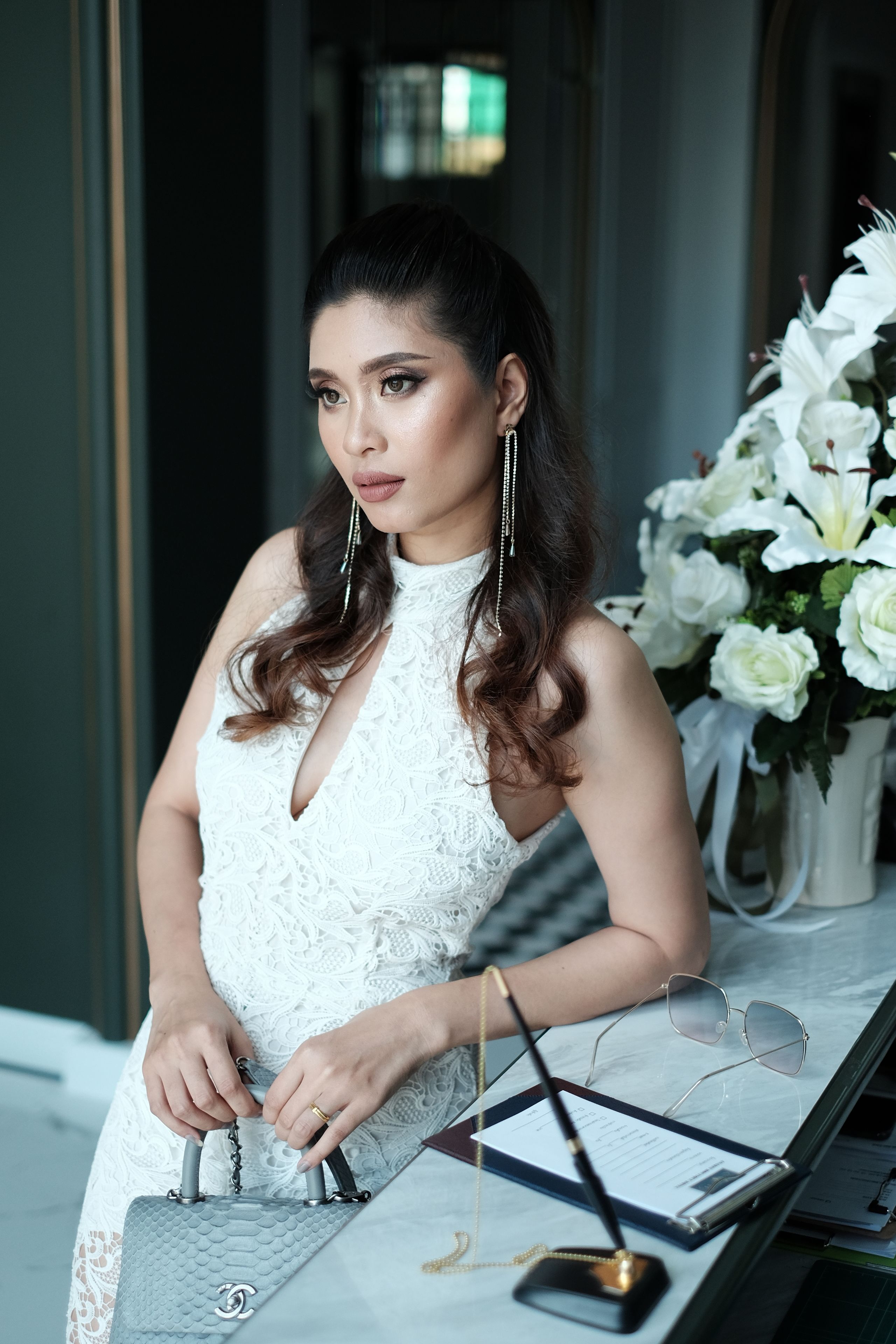 A woman in a white dress sitting at a table with a grey handbag, glasses, and a pen.