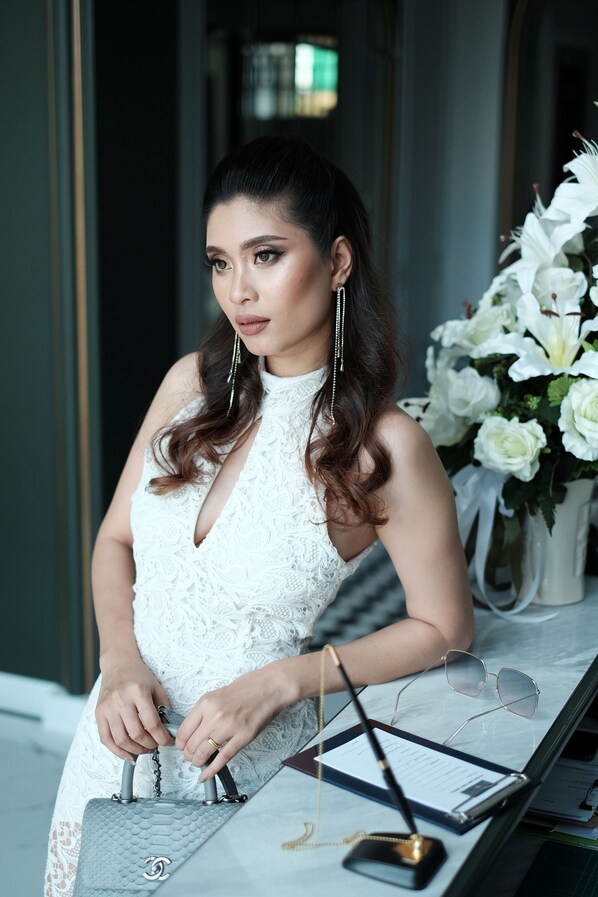 A woman in a white dress sitting at a table with a grey handbag, glasses, and a pen.