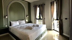 Uma cama bem arrumada com lençóis e toalhas brancas, uma grande janela em arco com cortinas e um aplique fixado na parede.