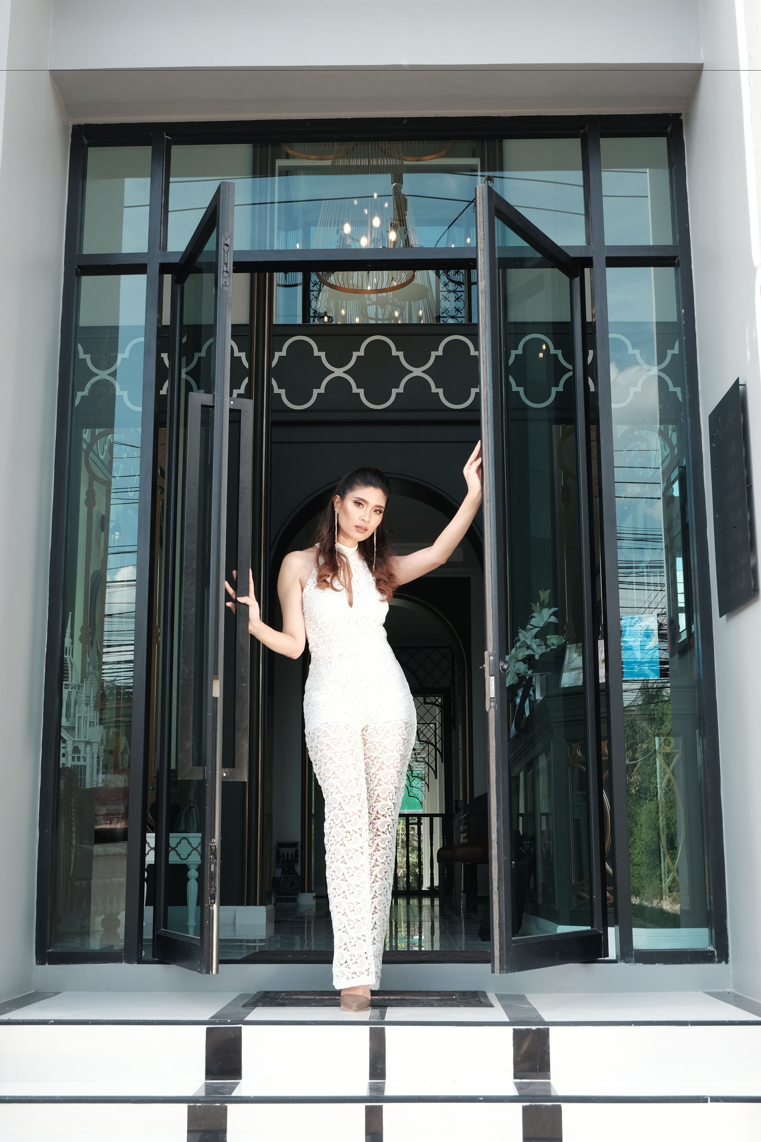 A woman in a white lace dress standing at the entrance of a building with large glass doors and a chandelier.