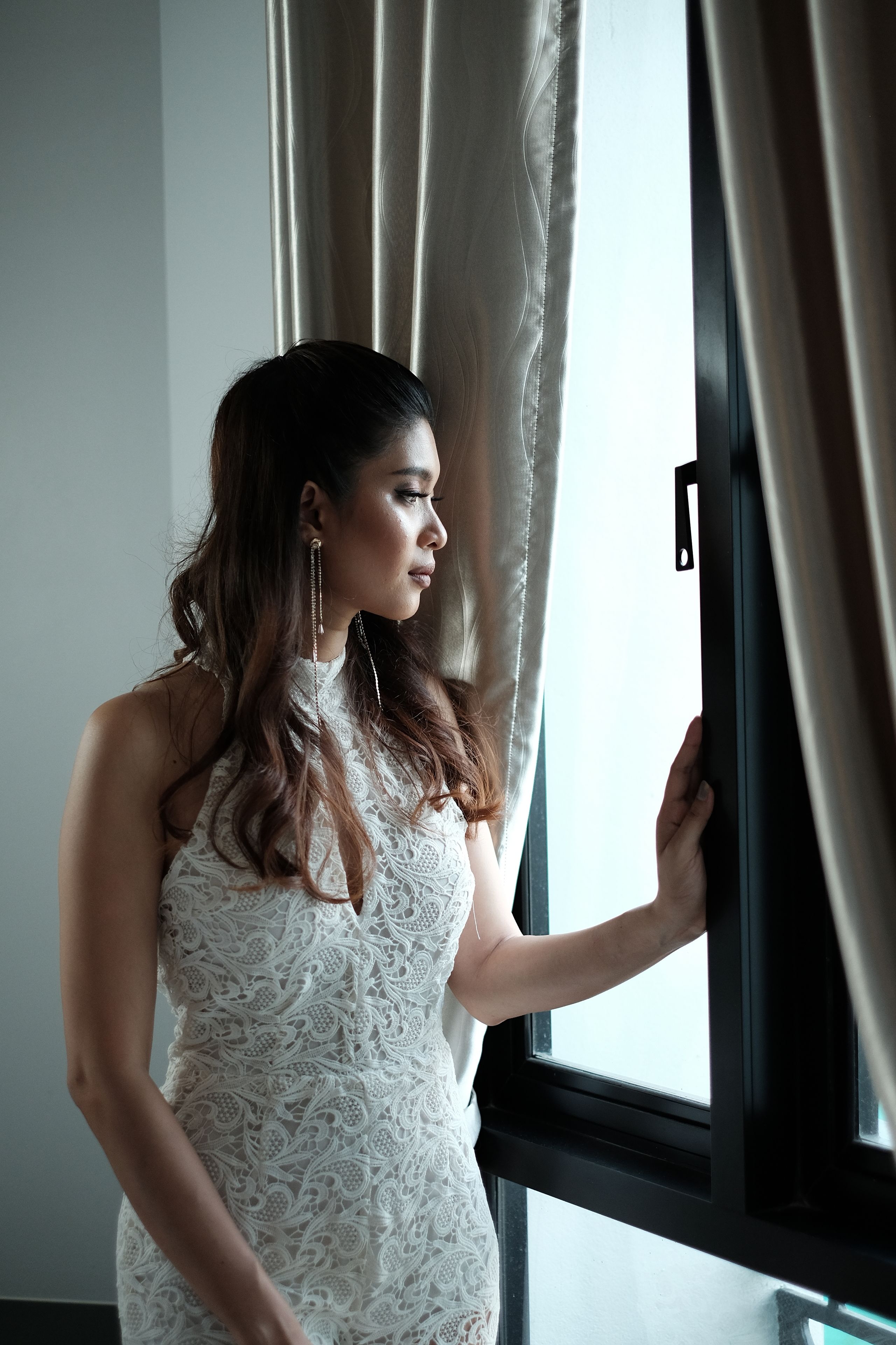 A person standing by a window with curtains, wearing a white lace dress.