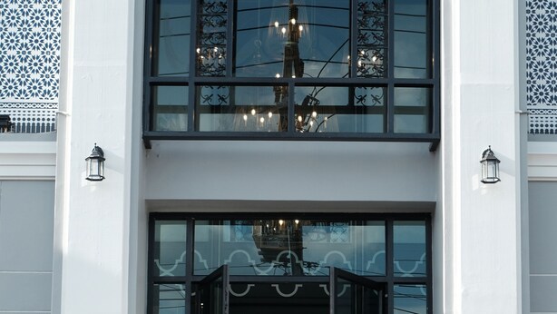 Mulher de vestido branco parada na entrada de um edifício de design moderno, com grandes portas de vidro e uma escadaria que leva à entrada.