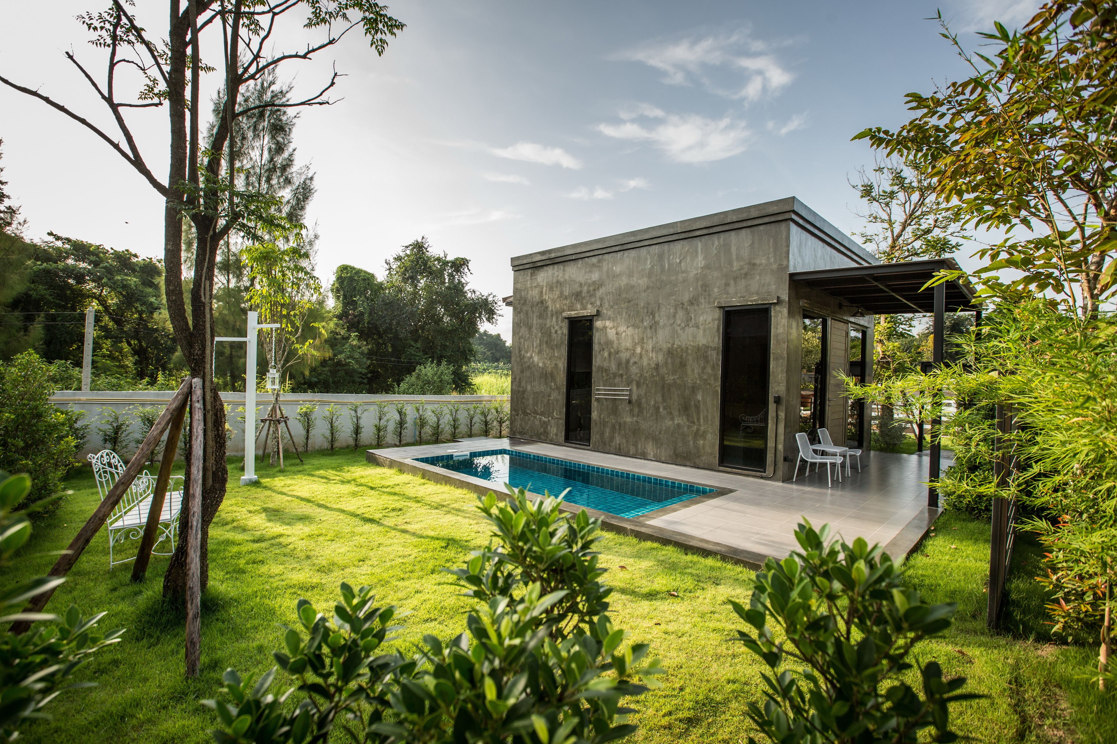 Una casa moderna con piscina, un patio con sillas y un jardín con árboles y arbustos.