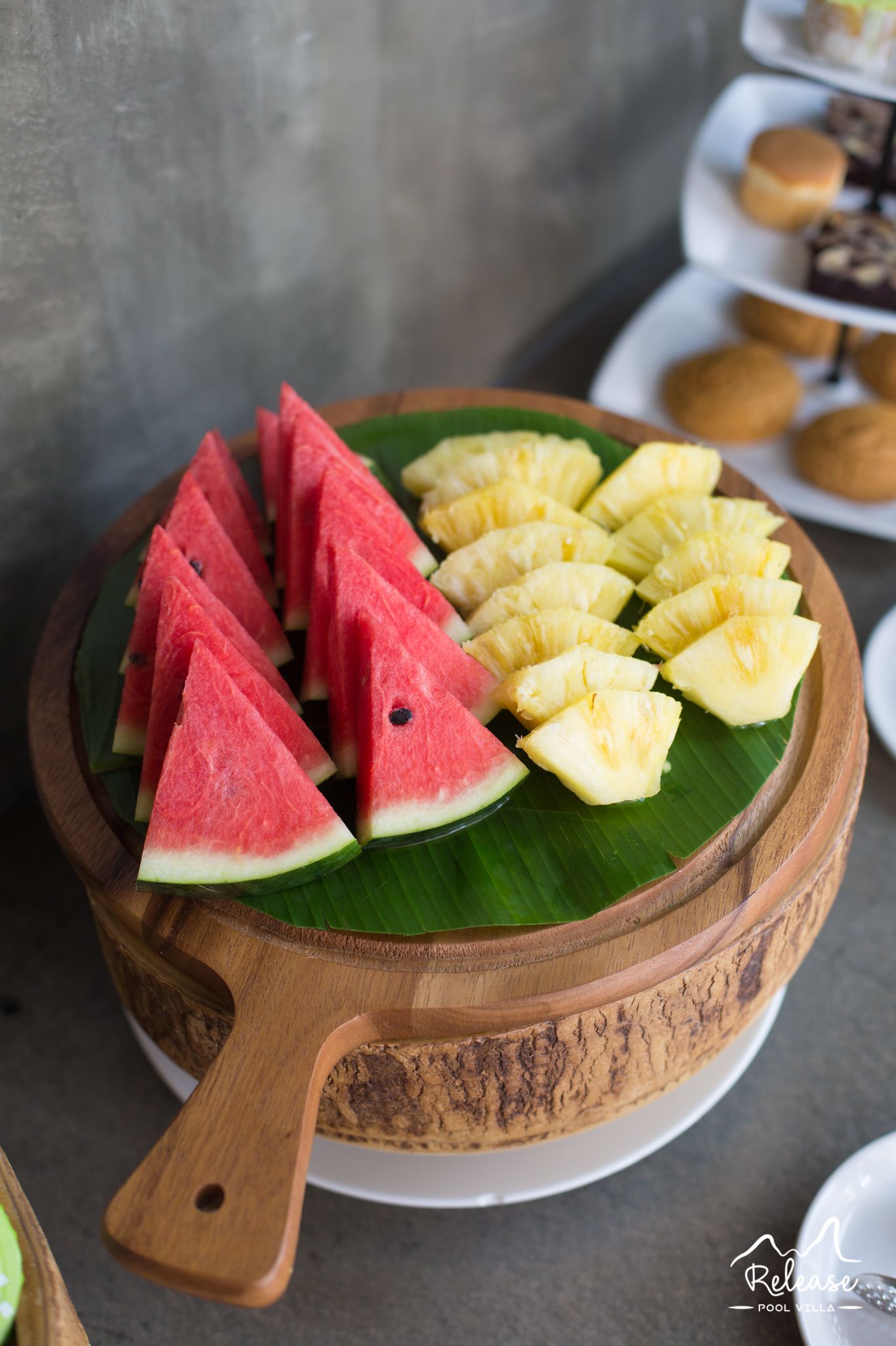 A wooden serving dish with sliced watermelon and pineapple on a banana leaf.