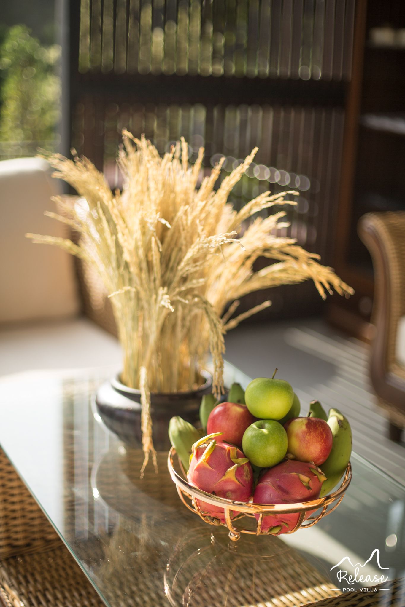 Une table en verre sur laquelle se trouve un panier en osier contenant des pommes et un vase avec des plantes séchées.