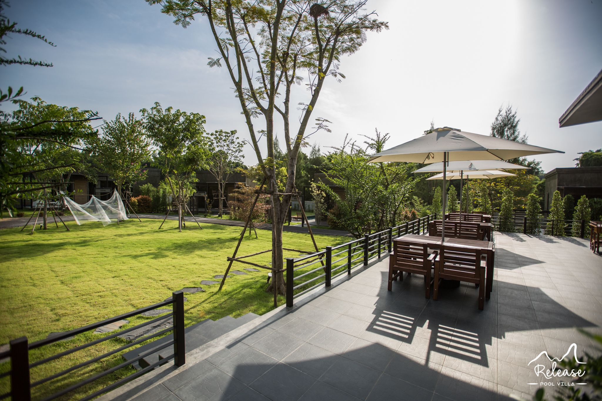 A patio area with a wooden table and chairs under a large umbrella, surrounded by a low fence and a grassy lawn.